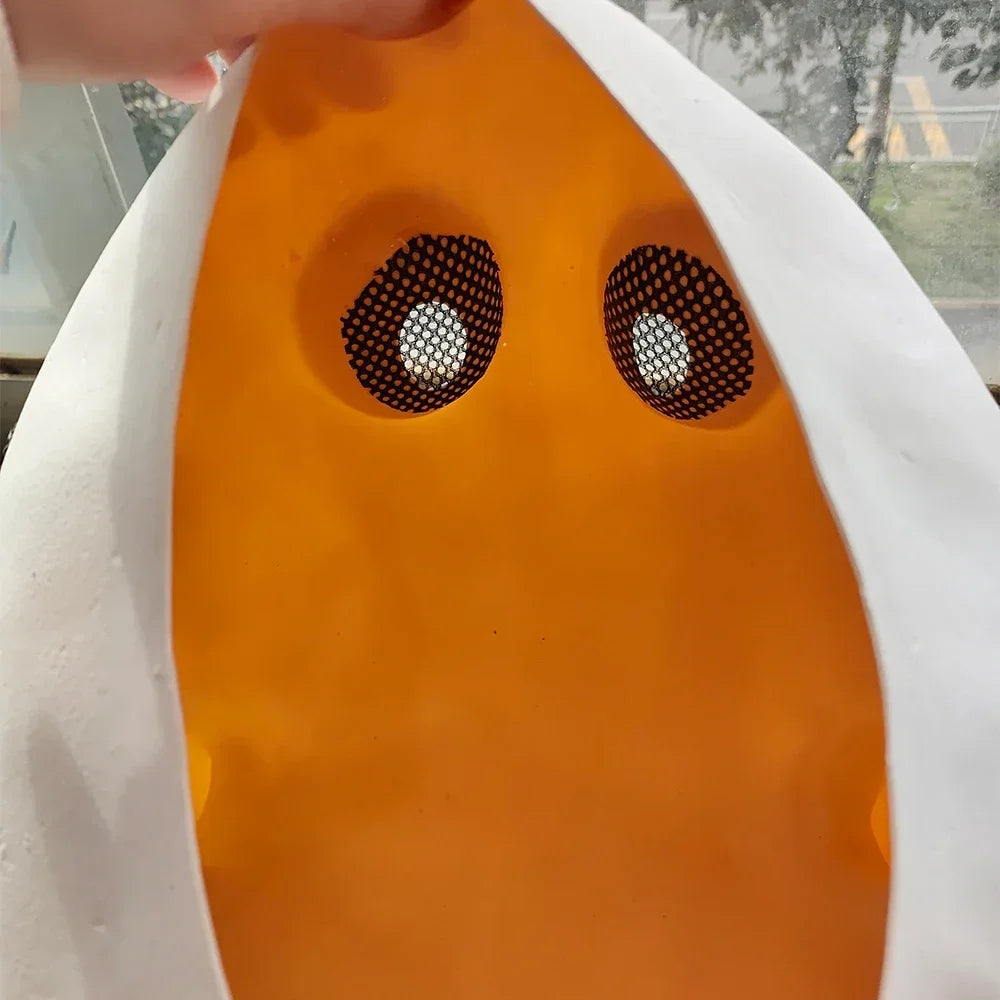 A close-up of a The Knight face mask or face cover with a bright orange center, black mesh eye holes, and white outer edges, being held open by a hand near the top.