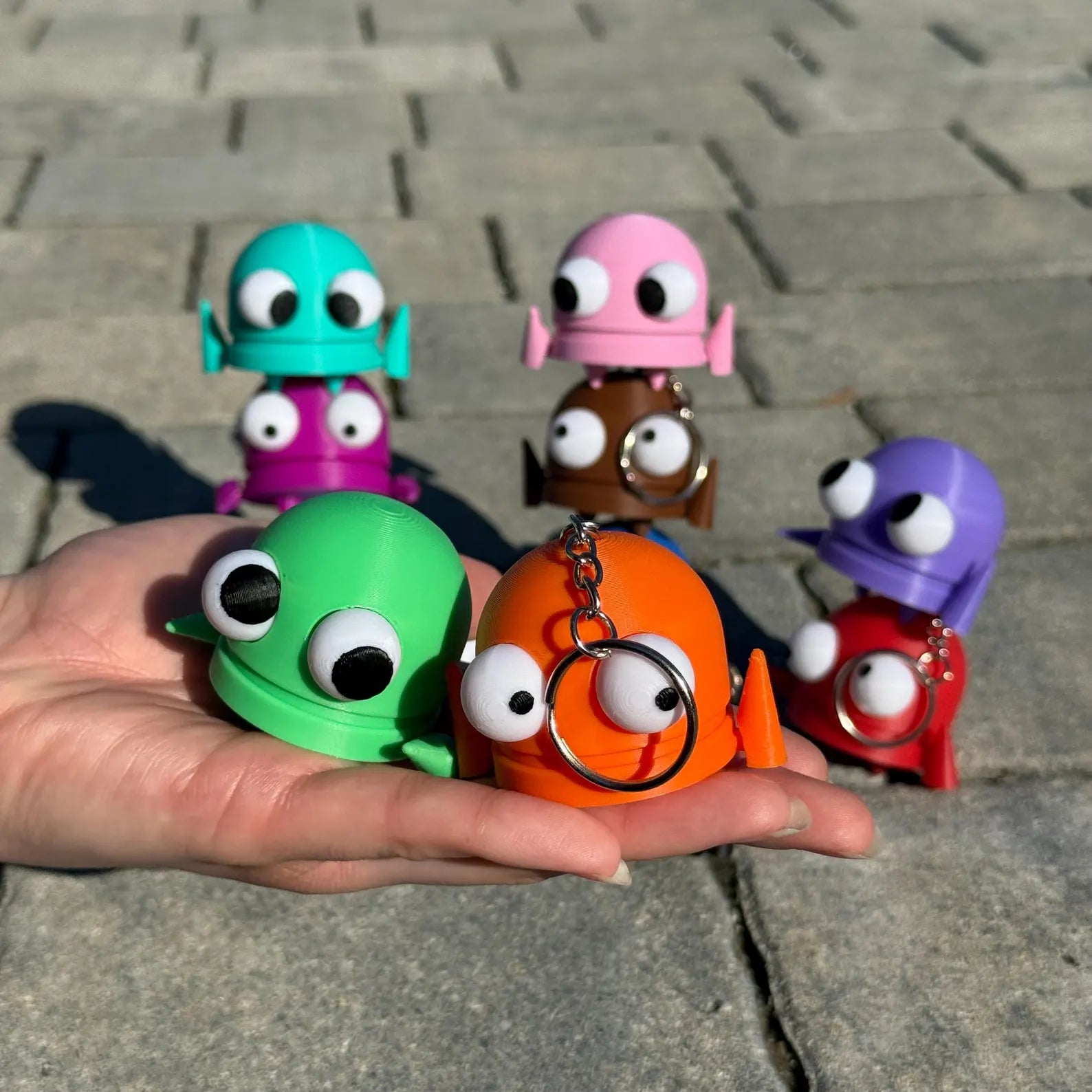 Colorful keychains shaped like R.E.P.O. characters with googly eyes held by a hand on a stone pavement.