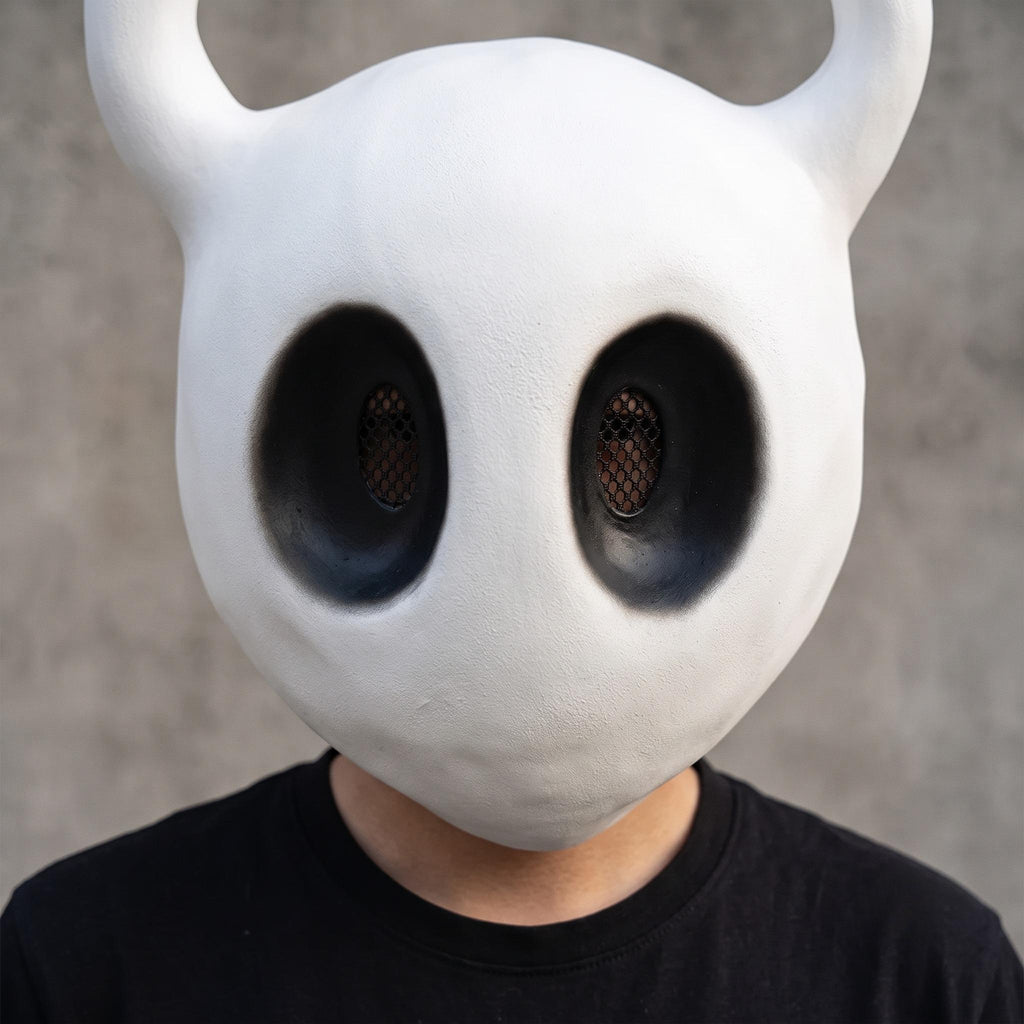 An adult cosplayer wearing the Hollow Knight mask paired with a traveler cloak for an authentic The Knight cosplay.