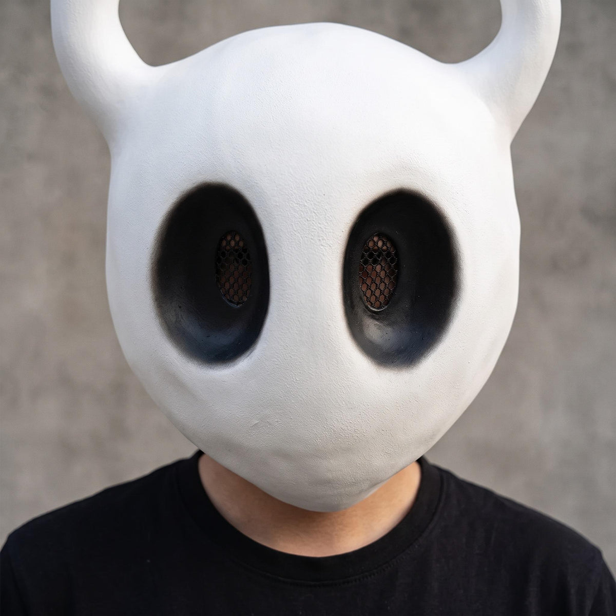 An adult cosplayer wearing the Hollow Knight mask paired with a traveler cloak for an authentic The Knight cosplay.