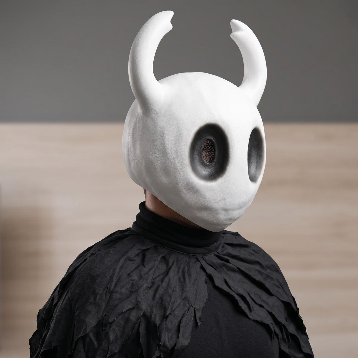 An adult cosplayer wearing the Hollow Knight mask paired with a traveler cloak for an authentic The Knight cosplay.